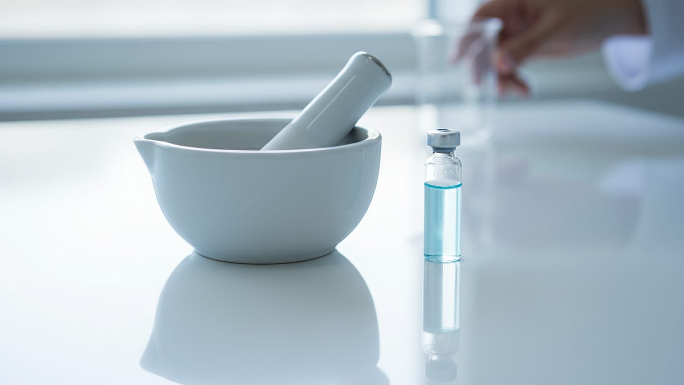 Illustration: A white mortar and pestle with a vial of pale blue liquid on a reflective white surface.