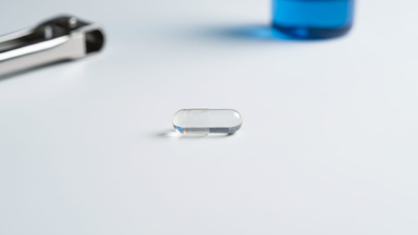 Illustration: An empty clear gelatin capsule on a white surface with soft, even lighting.