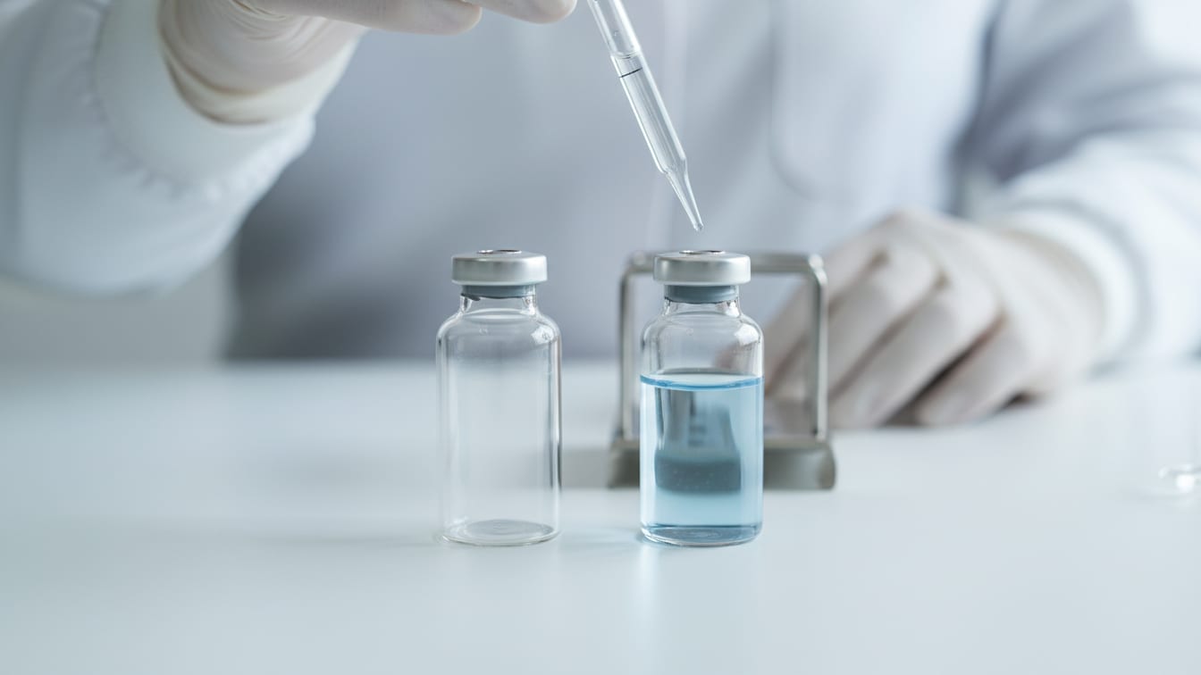 Illustration: Two clear glass vials, one with pale blue liquid, one empty, and a pipette on a white lab bench.